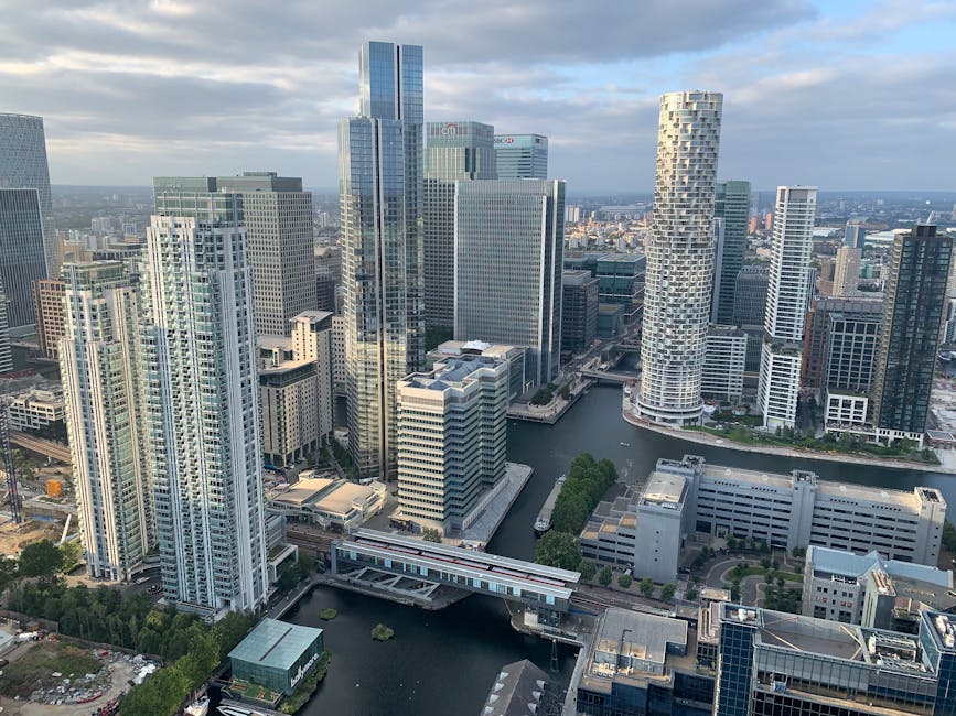 A wide-angle view of a modern city skyline featuring several high-rise buildings with varying architectural styles, situated along a waterway. In the foreground, a calm body of water reflects the surr