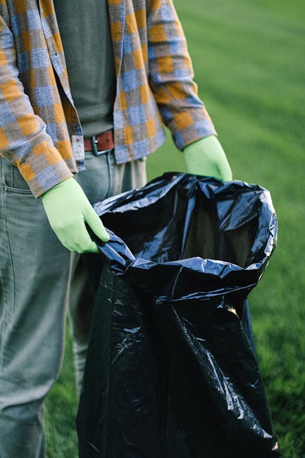A man and two children participating in beach cleanup activities during daylight hours, with the man holding a white trash bag in one hand and wearing casual clothing including a red t-shirt, denim sh