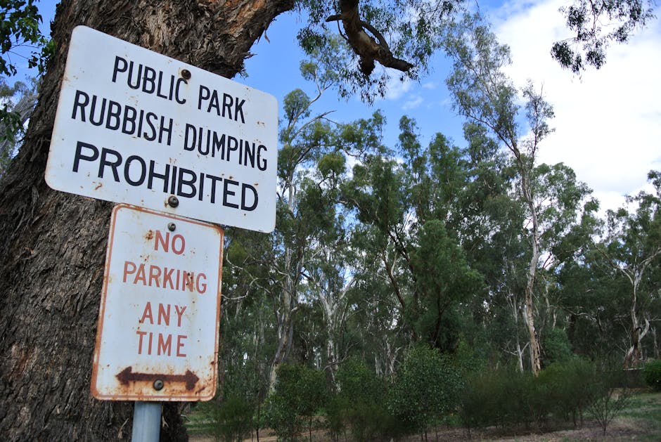 A white metal sign attached to a tree in a wooded area, with the top sign reading 'PUBLIC PARK RUBBISH DUMPING PROHIBITED' in bold black letters, and the lower sign stating 'NO PARKING ANY TIME' in re